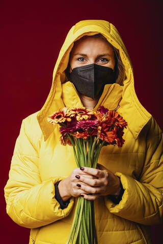 Woman wearing face mask and hooded shirt holding flower while standing against red background stock photo