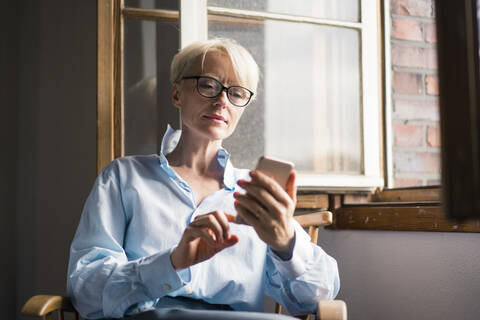 Mature businesswoman using mobile phone while sitting on chair at window in home office stock photo