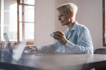 Lächelnde Geschäftsfrau, die eine Kaffeetasse in der Hand hält, während sie an einem Laptop am Schreibtisch im Heimbüro arbeitet - MOEF03577