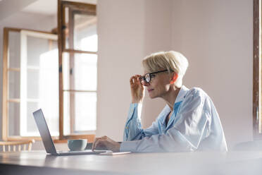 Geschäftsfrau passt ihre Brille an, während sie zu Hause am Schreibtisch am Laptop arbeitet - MOEF03579