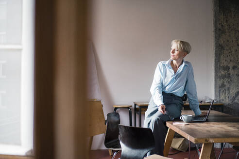 Lächelnde Geschäftsfrau mit Laptop auf dem Schreibtisch sitzend, während sie im Büro zu Hause wegschaut - MOEF03581
