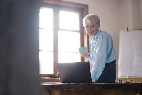 Lächelnde Geschäftsfrau, die eine Kaffeetasse in der Hand hält, während sie einen Laptop auf dem Schreibtisch im Heimbüro benutzt - MOEF03582