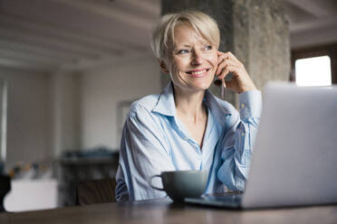 Smiling businesswoman with laptop and coffee cup looking away while talking on mobile phone at desk in home office - MOEF03593