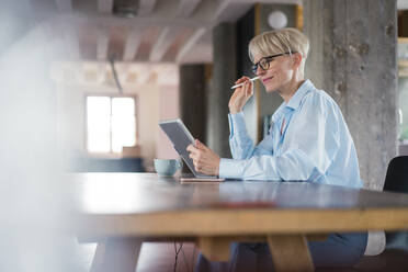 Lächelnde Geschäftsfrau, die ein digitales Tablet benutzt, während sie am Schreibtisch im Heimbüro sitzt - MOEF03594