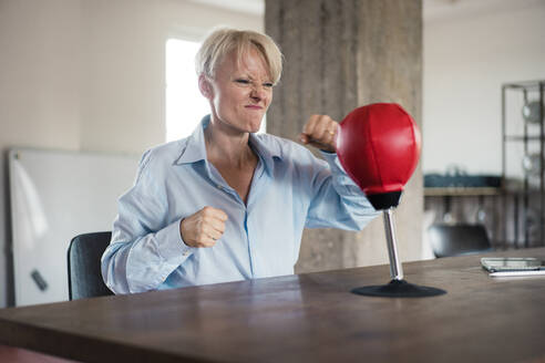 Blond businesswoman punching boxing bag on table at home office - MOEF03595