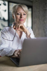Lächelnde Unternehmerin mit Brille bei der Arbeit am Laptop im Home Office - MOEF03599