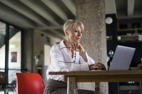 Lächelnde Geschäftsfrau, die eine Brille hält, während sie einen Laptop am Schreibtisch neben einer Säule im Büro zu Hause benutzt, lizenzfreies Stockfoto