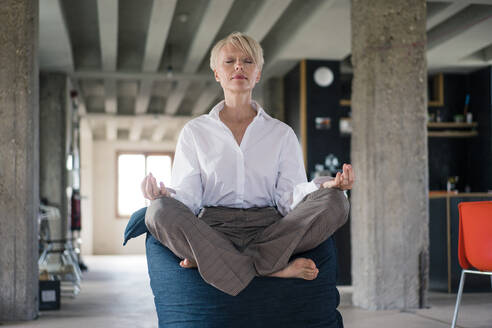 Blonde Geschäftsfrau meditiert, während sie zu Hause auf einem Sitzsack sitzt - MOEF03607