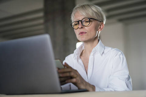Businesswoman with laptop using smart phone while sitting at home office stock photo