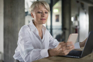 Blond businesswoman with laptop using mobile phone while sitting at desk in home office - MOEF03614