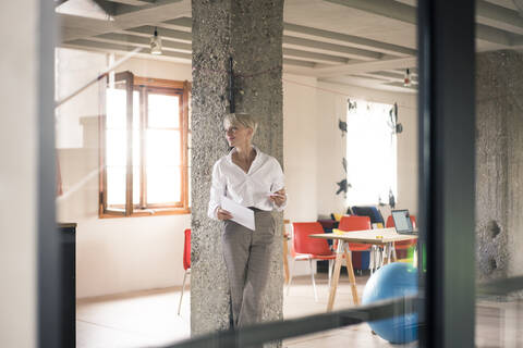 Geschäftsfrau durch Glaswand gesehen, während sie ein Dokument gegen eine Säule in ihrem Büro hält, lizenzfreies Stockfoto