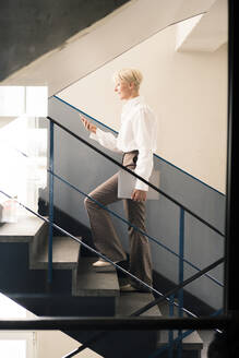 Weibliche Unternehmerin, die ein Smartphone benutzt, während sie auf einer Treppe in einer Loftwohnung geht - MOEF03623