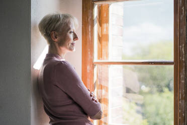 Lächelnde Geschäftsfrau, die durch ein Fenster schaut und sich an die Wand lehnt - MOEF03627