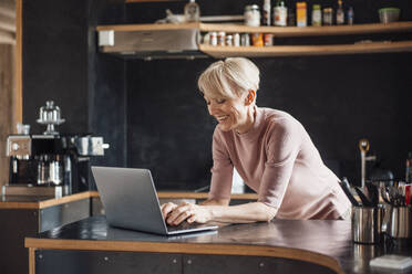 Glückliche Geschäftsfrau arbeitet am Laptop an der Küchentheke im Heimbüro - MOEF03630
