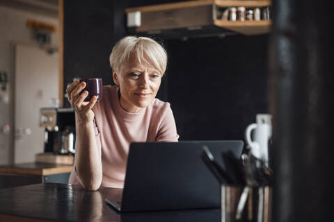 Lächelnde Geschäftsfrau, die auf einen Laptop schaut, während sie eine Kaffeetasse am Küchentisch im Heimbüro hält, lizenzfreies Stockfoto