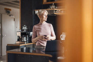 Lächelnde Geschäftsfrau, die wegschaut, während sie eine Kaffeetasse in der Küche ihres Heimbüros hält - MOEF03632