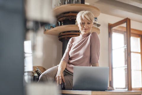 Female freelance worker with laptop sitting on kitchen counter at home stock photo