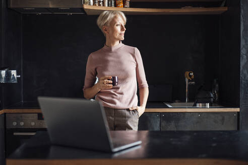 Female entrepreneur with hands in pockets looking away while having coffee in kitchen in home office - MOEF03634