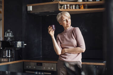 Smiling businesswoman looking away while holding coffee cup in kitchen at home - MOEF03635