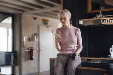 Smiling businesswoman with coffee cup using mobile phone while sitting on kitchen counter at home - MOEF03636