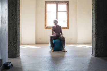 Businesswoman using smart phone while sitting on fitness ball in loft apartment - MOEF03638