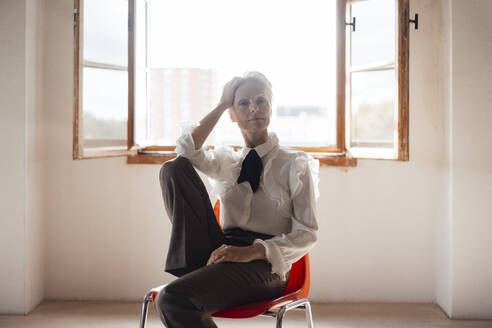 Seriöse Geschäftsfrau mit Hand in den Haaren, die auf einem Stuhl vor dem Fenster in einem Büro zu Hause sitzt - MOEF03641