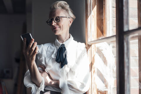 Smiling businesswoman looking at mobile phone while standing at window in home office stock photo