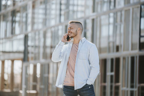 Smiling man talking on mobile phone while standing with hands in pockets outdoors stock photo