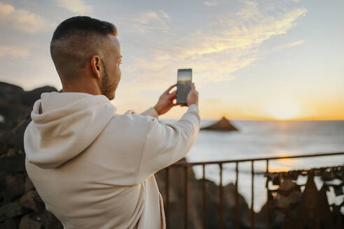 Mann fotografiert den Sonnenaufgang über dem Meer mit seinem Mobiltelefon, während er auf einer Brücke steht - MIMFF00566