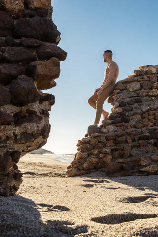 Mann ohne Hemd schaut weg, während er sich an eine Steinmauer am Strand lehnt, lizenzfreies Stockfoto
