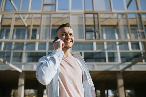 Mittlerer erwachsener Mann, der lächelt, während er mit einem Mobiltelefon spricht, das an einem Gebäude steht, lizenzfreies Stockfoto