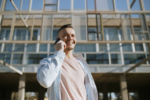 Mid adult man smiling while talking on mobile phone standing against building - MIMFF00575