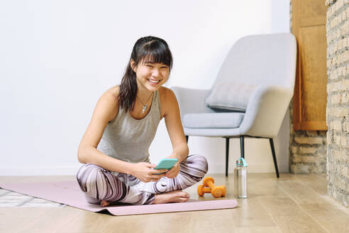 Lächelnde Frau, die ein Smartphone benutzt, während sie zu Hause im Schneidersitz sitzt - AODF00316