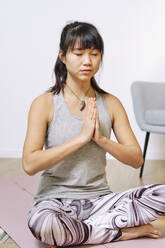 Young woman sitting with hands clasped while practicing yoga at home - AODF00318