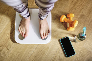 Woman checking weight while standing on weight scale by dumbbell, water bottle and mobile phone at home - AODF00325