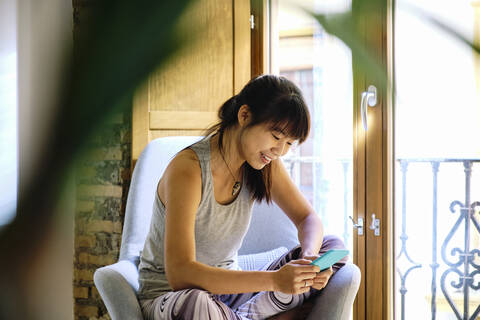 Lächelnde Frau, die ein Mobiltelefon benutzt, während sie auf einem Stuhl zu Hause sitzt, lizenzfreies Stockfoto