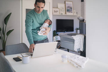 Businessman carrying baby boy while working on laptop at home office - MPPF01526