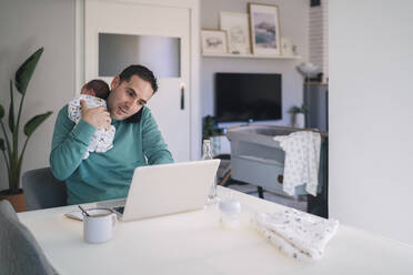 Man with baby boy talking on mobile phone while using laptop at desk in home office - MPPF01528