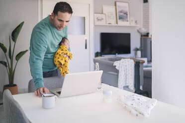 Father carrying son while looking at laptop in home office - MPPF01532
