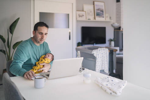 Single father working on laptop while carrying son at desk in home office - MPPF01535