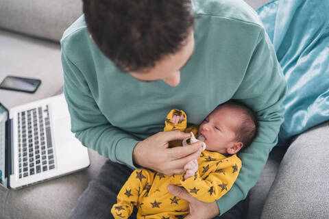 Vater steckt Schnuller in den Mund seines Sohnes, während er zu Hause am Laptop sitzt, lizenzfreies Stockfoto