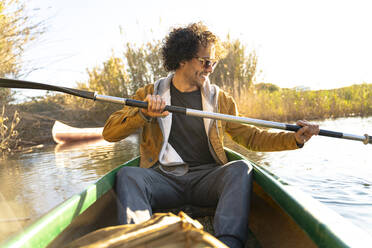 Smiling man wearing sunglasses paddling through oar while sitting in canoe on river - SBOF02677