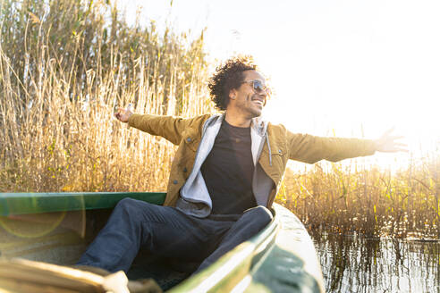 Carefree man with arms outstretched smiling while sitting in canoe on river - SBOF02683