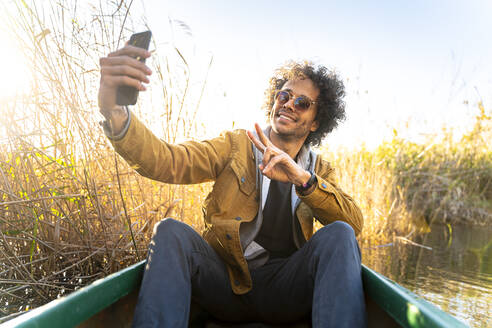 Young man gesturing peace sign while taking selfie through mobile phone sitting in canoe on river - SBOF02684