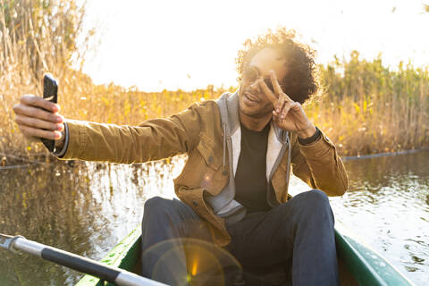Junger Mann zeigt Friedenszeichen, während er ein Selfie mit seinem Handy macht, während er in einem Kanu auf einem Fluss sitzt, lizenzfreies Stockfoto