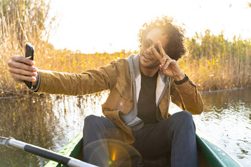 Junger Mann zeigt Friedenszeichen, während er ein Selfie mit seinem Handy macht, während er in einem Kanu auf einem Fluss sitzt - SBOF02687