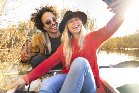 Lächelnde Frau, die ein Selfie mit einem Mann durch ein Mobiltelefon macht, während sie in einem Kanu auf einem Fluss sitzt, lizenzfreies Stockfoto