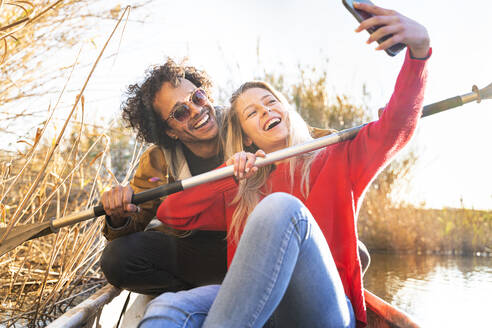 Happy couple taking selfie with oar through mobile phone while sitting in canoe on river - SBOF02693