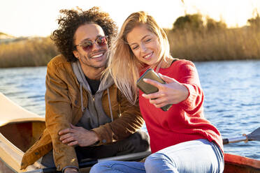 Lächelndes Paar, das ein Selfie mit dem Mobiltelefon macht, während es in einem Kanu auf einem Fluss sitzt - SBOF02695