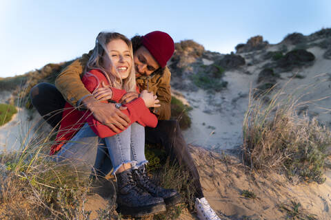 Affectionate couple smiling while sitting on sand dune during sunset stock photo